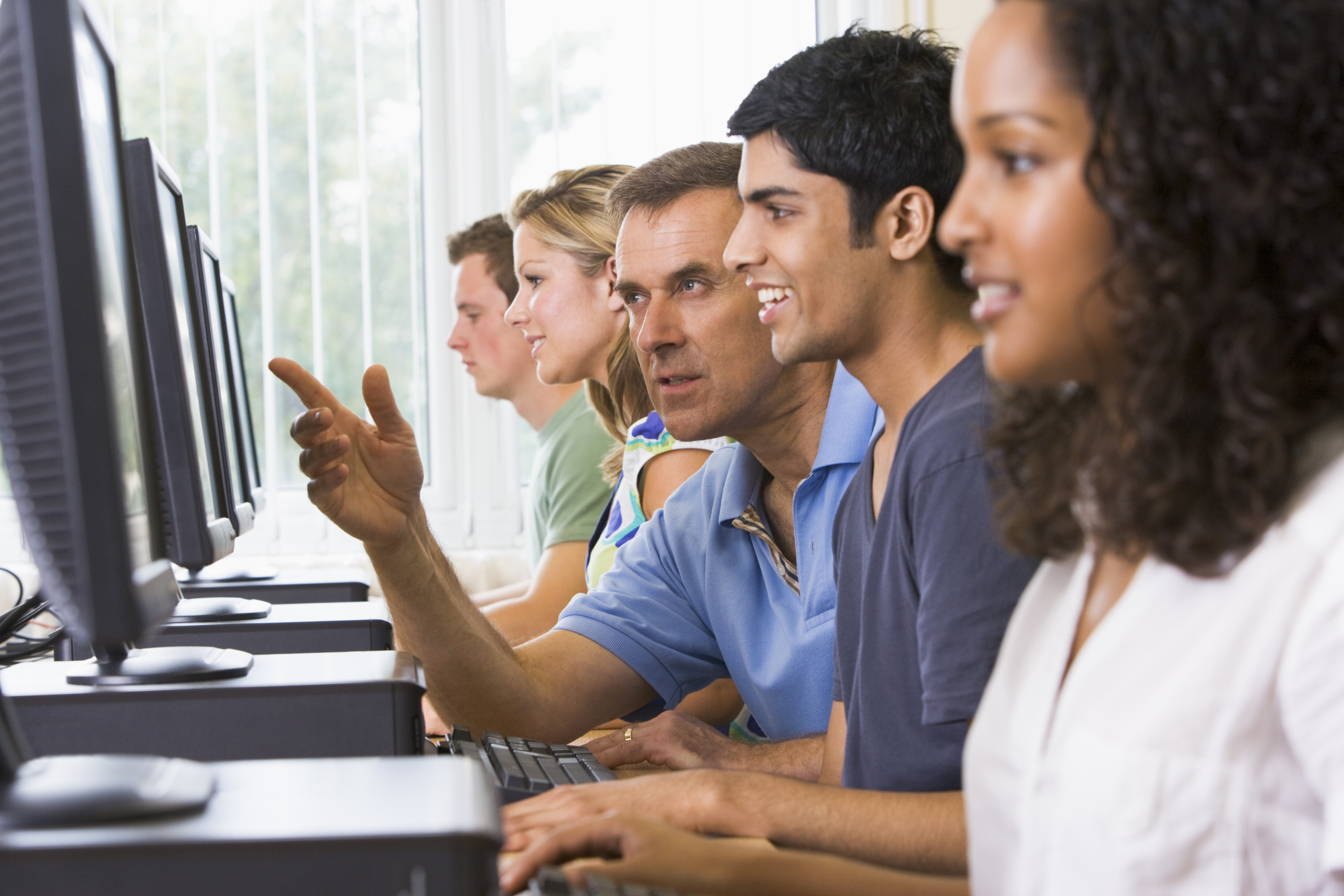 A imagem mostra homens e mulheres utilizando computadores com ajuda de um professor.