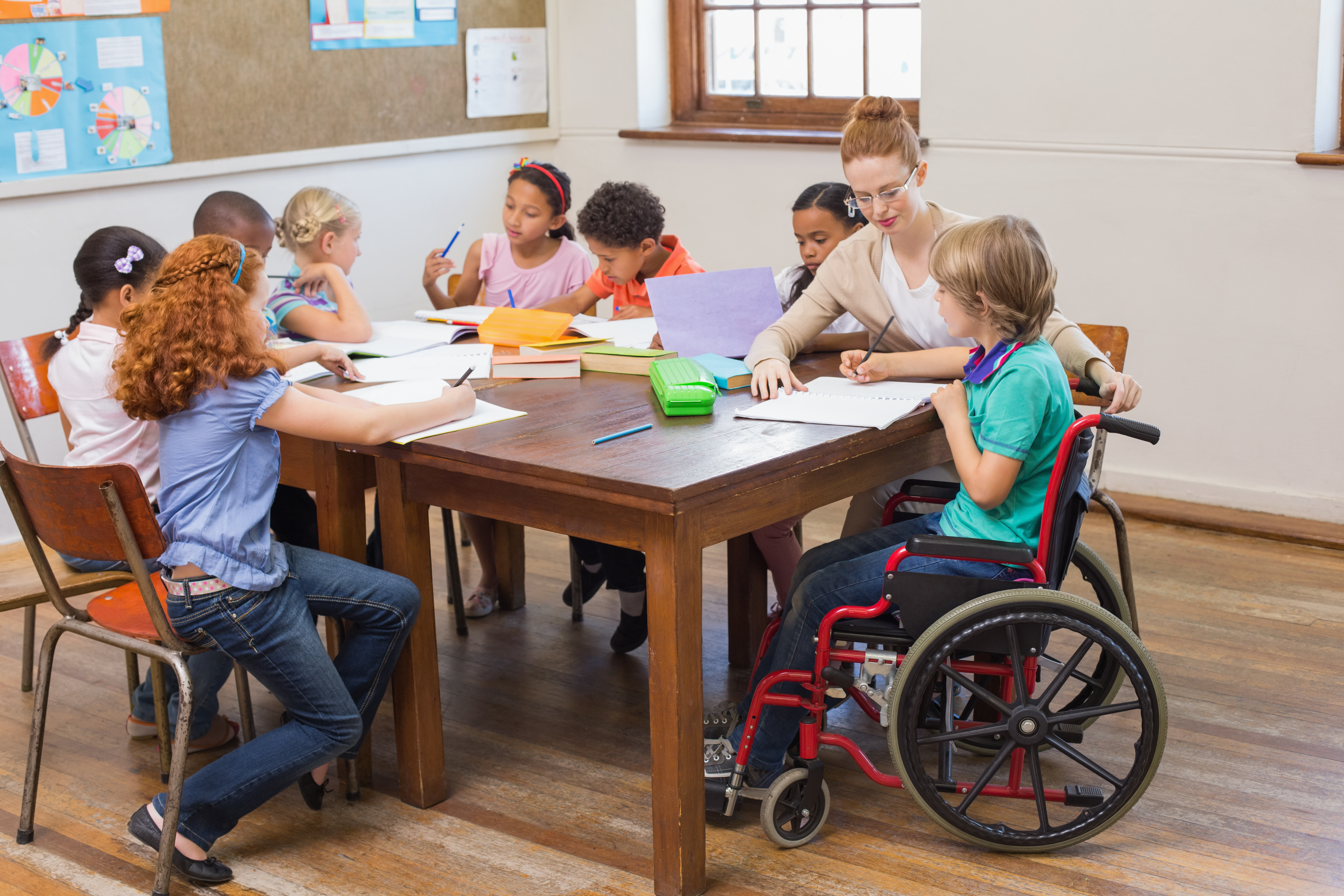 A imagem mostra uma sala de aula com alunos sentados ao redor de uma mesa, interagindo com a professora.