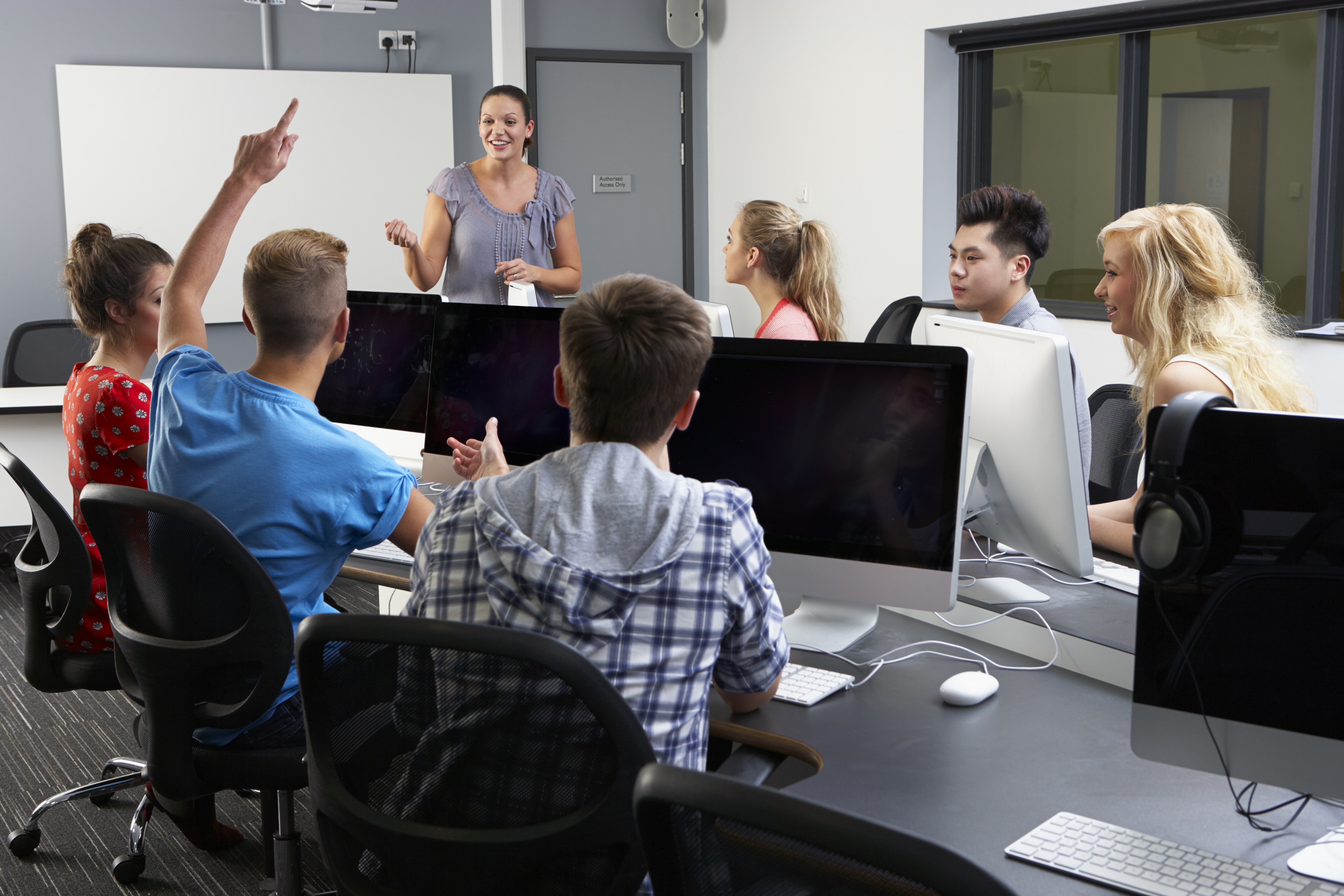 A imagem mostra uma professora em sala de aula com alunos.